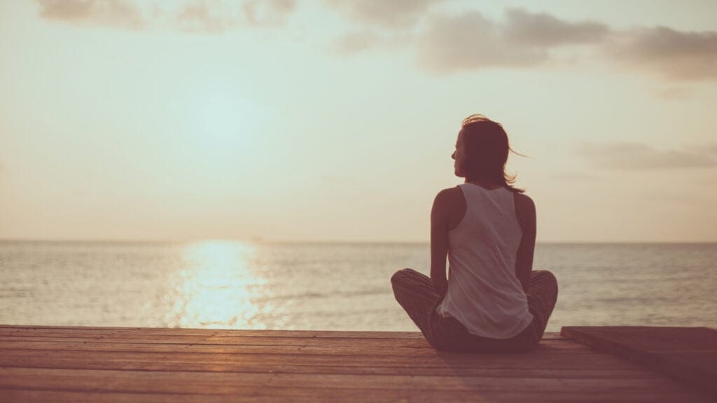 Woman sitting cross-legged on a wooden deck, peacefully watching the sunset over the ocean.