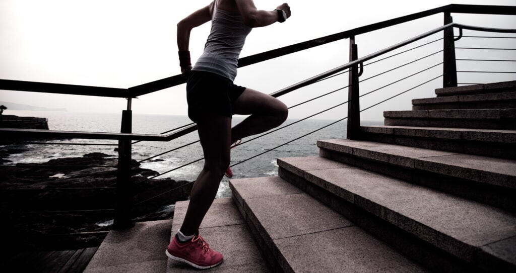 Woman running up outdoor stairs by the sea, wearing athletic gear and pink shoes.