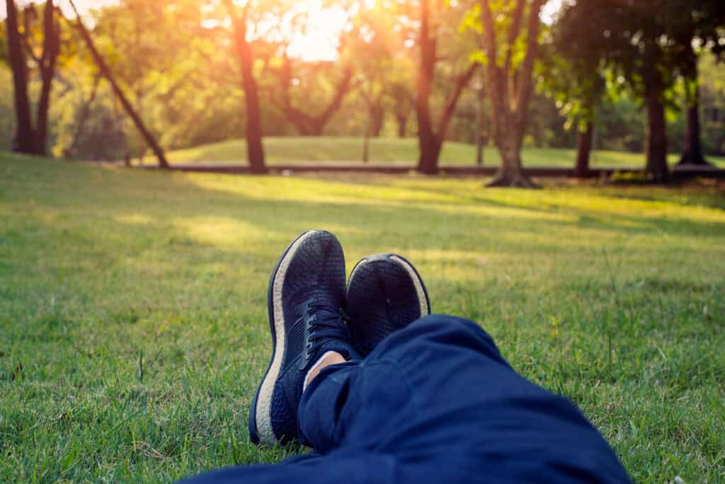 Person lying on grass in a sunlit park, wearing navy sneakers and pants, relaxing under trees.