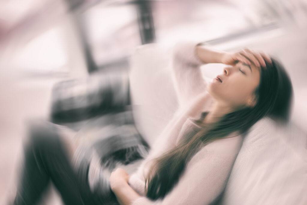 A woman sitting on a couch with a blurred, dizzy background effect, holding her head and stomach, appearing exhausted and overwhelmed - representing chronic fatigue or burnout