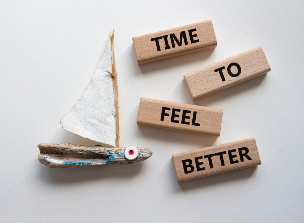Wooden sailboat decoration next to wooden blocks with the words "TIME TO FEEL BETTER" arranged on a light background.