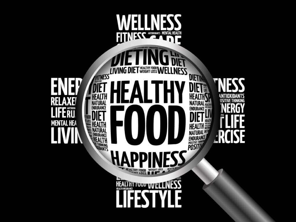 A magnifying glass highlighting the words “HEALTHY FOOD” in the centre of a collage of health-related terms such as “DIET,” “WELLNESS,” “HAPPINESS,” and “LIFESTYLE” on a black background