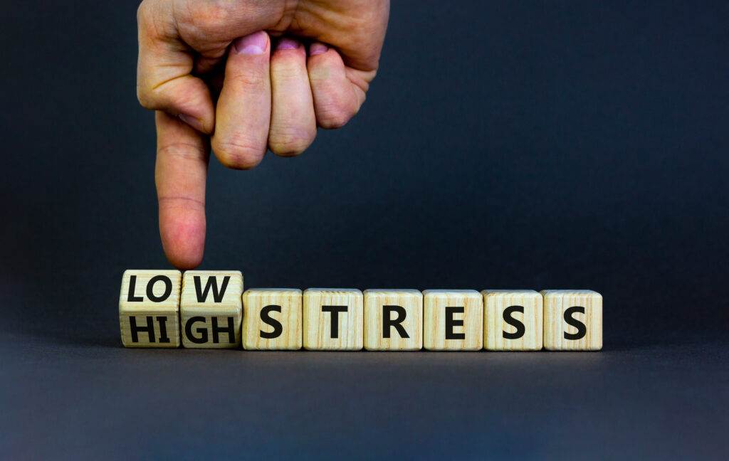 Hand flipping a wooden block between the words “HIGH” and “LOW” in front of the word “STRESS,” symbolising a shift in stress levels against a dark background