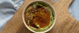 A bowl of green hummus topped with a drizzle of olive oil and sprinkled with mixed seeds and spices sits on a wooden board, with a light blue cloth partially visible in the background.