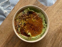 A bowl of green hummus topped with a drizzle of olive oil and sprinkled with mixed seeds and spices sits on a wooden board, with a light blue cloth partially visible in the background.