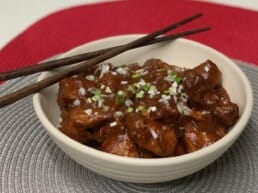 A bowl of dark, saucy chicken garnished with chopped green onions and sesame seeds, with a pair of chopsticks resting on top. The bowl is set on a gray and red placemat.