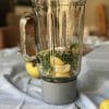 A blender filled with spinach, lemon slices, and other ingredients sits on a blue cloth with whole lemons nearby.