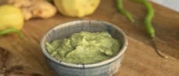 A small bowl filled with green chutney sits on a wooden surface, with fresh green chilies, ginger, and a lemon in the background.