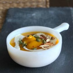White ceramic bowl filled with creamy butternut squash soup, garnished with a swirl of fresh cream, slivered almonds, pumpkin seeds, and a sprinkle of red chilli flakes.