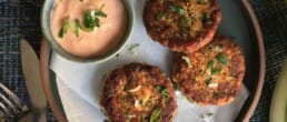 Three golden-brown patties garnished with herbs are served on a plate with a small bowl of creamy dipping sauce topped with fresh green herbs. A fork and knife rest nearby on the table.