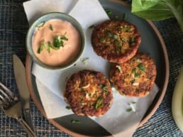 Three golden-brown patties garnished with herbs are served on a plate with a small bowl of creamy dipping sauce topped with fresh green herbs. A fork and knife rest nearby on the table.