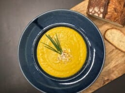 A bowl of smooth carrot and orange soup served in a deep blue ceramic plate. The bowl is placed on a rustic wooden board alongside a slice of toasted sourdough and a loaf of crusty bread. The background is a textured white surface, creating a cozy, artisanal presentation.