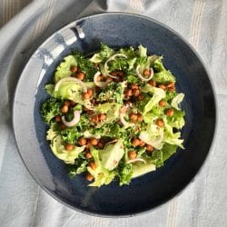 A dark ceramic plate filled with a vibrant kale and chickpea salad, topped with onion rings, sprinkled seeds, and a light dressing, placed on a soft blue cloth.