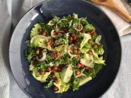 A vibrant salad made of kale, romaine lettuce, roasted chickpeas, and slivered red onions, garnished with sesame seeds and chilli flakes. A light cashew dressing is drizzled over the greens.
