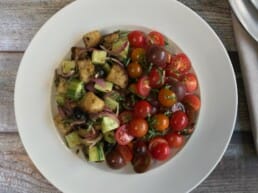 A white plate holds a colorful salad with cherry tomatoes, cucumber, red onion, croutons, and olives. Wooden salad servers rest on a white napkin beside the plate on a light stone surface.