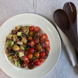 A white plate holds a colorful salad with cherry tomatoes, cucumber, red onion, croutons, and olives. Wooden salad servers rest on a white napkin beside the plate on a light stone surface.
