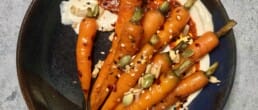 A dark ceramic plate featuring glazed baby carrots arranged over a swirl of creamy harissa-spiked sauce, garnished with mixed seeds including pumpkin, sesame, and sunflower, placed on a grey stone surface.