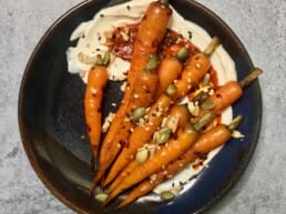A dark ceramic plate featuring glazed baby carrots arranged over a swirl of creamy harissa-spiked sauce, garnished with mixed seeds including pumpkin, sesame, and sunflower, placed on a grey stone surface.