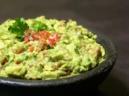 A close-up of a bowl of guacamole garnished with chopped tomatoes and a sprig of cilantro on top, with a dark background.