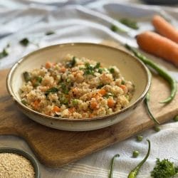 A bowl of colourful quinoa upma garnished with fresh coriander, featuring finely chopped carrots, green peas, and French beans. The dish is served in a rustic ceramic bowl placed on a wooden board, surrounded by raw green chilies, whole quinoa grains, and fresh carrots in the background. The scene is softly lit with a casual, homely table setting.