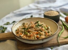 A close-up of a rustic ceramic bowl filled with quinoa upma, featuring vibrant chopped carrots, green peas, French beans, and garnished with fresh coriander. The bowl rests on a wooden board, surrounded by green chilies, raw quinoa in a small bowl, and fresh vegetables, creating a warm and inviting food presentation.