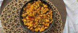 A bowl of scrambled paneer mixed with vegetables sits on a woven placemat, viewed from above. The dish is vibrant with orange, yellow, and green colors.