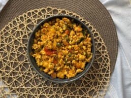 A bowl of scrambled paneer mixed with vegetables sits on a woven placemat, viewed from above. The dish is vibrant with orange, yellow, and green colors.