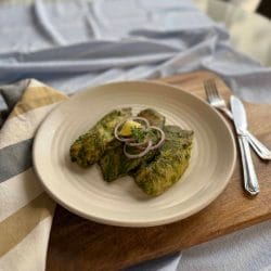 Three pieces of green vegetable fritters are served on a beige plate, garnished with sliced onions and a lemon wedge, placed on a wooden board with a fork, knife, and a striped napkin nearby.