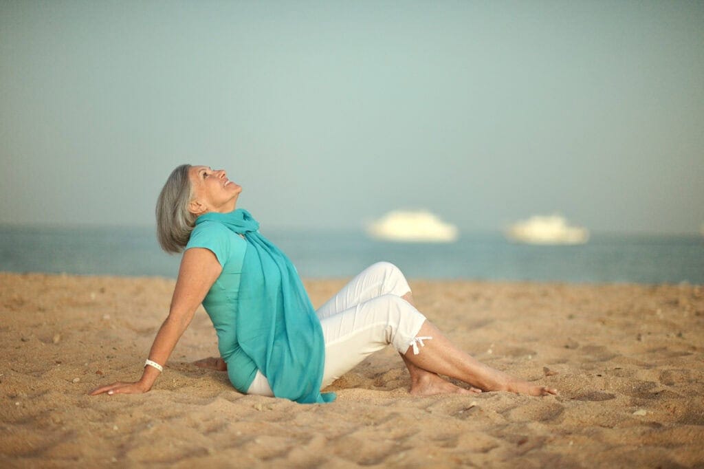 An older woman sitting relaxed on a sandy beach, leaning back and looking up peacefully, with the ocean and blurred boats in the background.