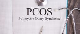 Open notebook with the words &ldquo;PCOS Polycystic Ovary Syndrome&rdquo; written on a grid page, accompanied by a stethoscope and metal pen on a wooden surface