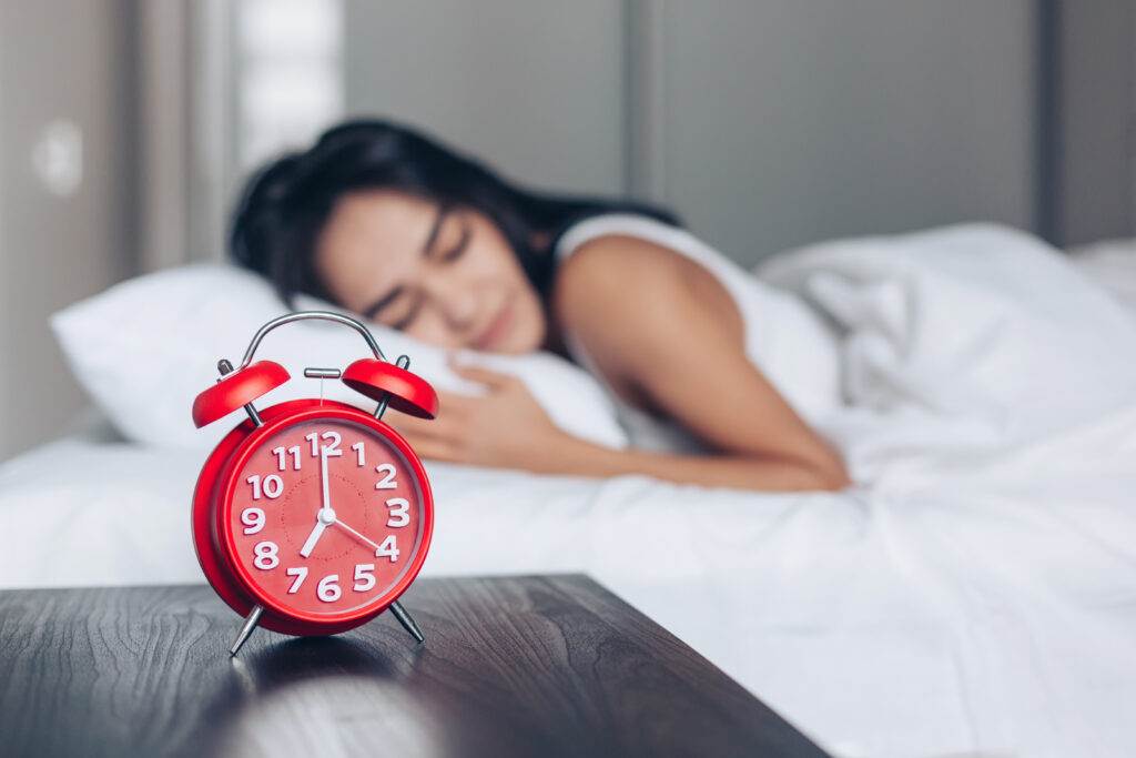 A red alarm clock in focus on a bedside table with a woman peacefully sleeping in the background - symbolising the importance of sleep routines and restful habits