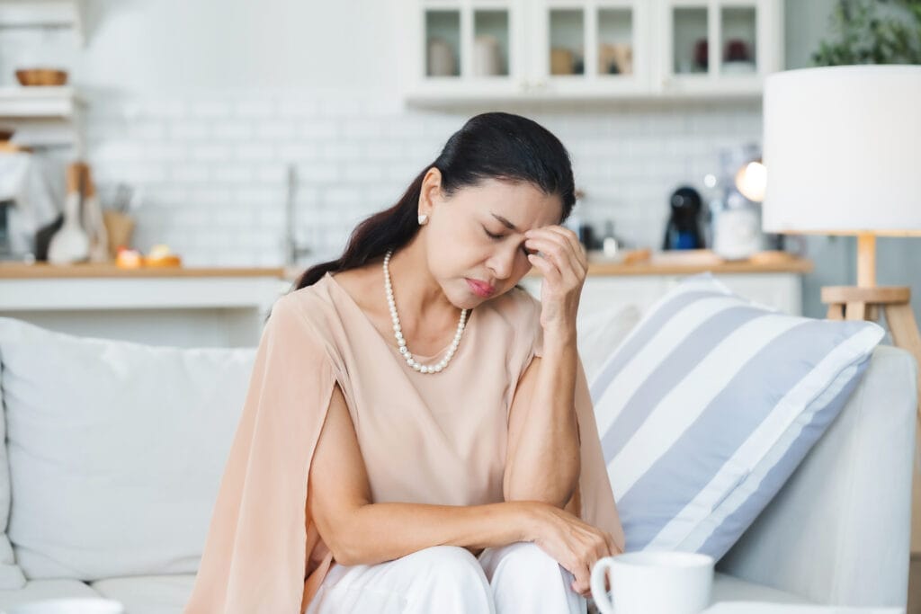A middle-aged woman sitting on a couch with her eyes closed and hand on her forehead, appearing stressed or anxious, in a modern kitchen setting.
