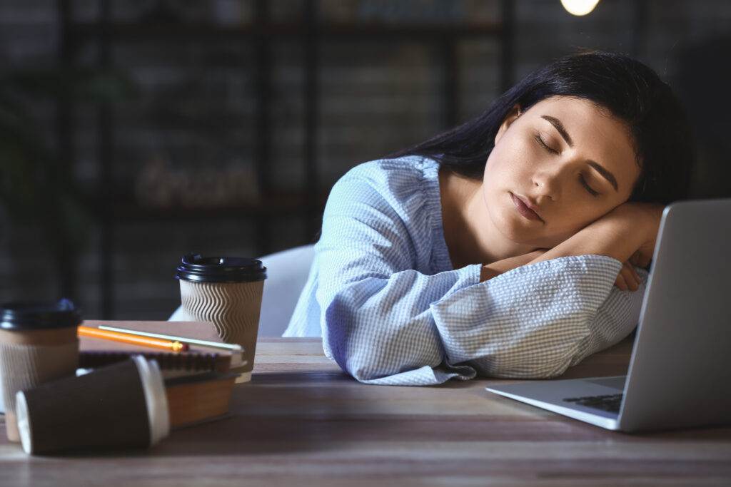 Tired woman in a checkered blue shirt sleeping at a desk beside a laptop, surrounded by takeaway coffee cups, notebooks, and a pencil