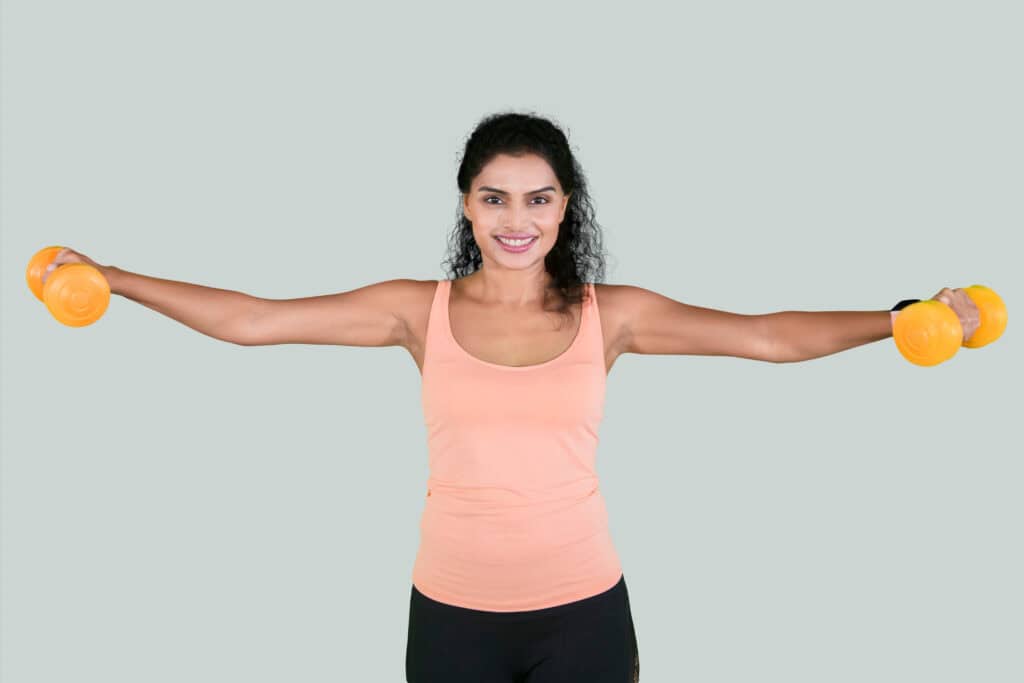 Woman in workout attire holding yellow dumbbells with arms extended sideways, demonstrating a lateral raise exercise.