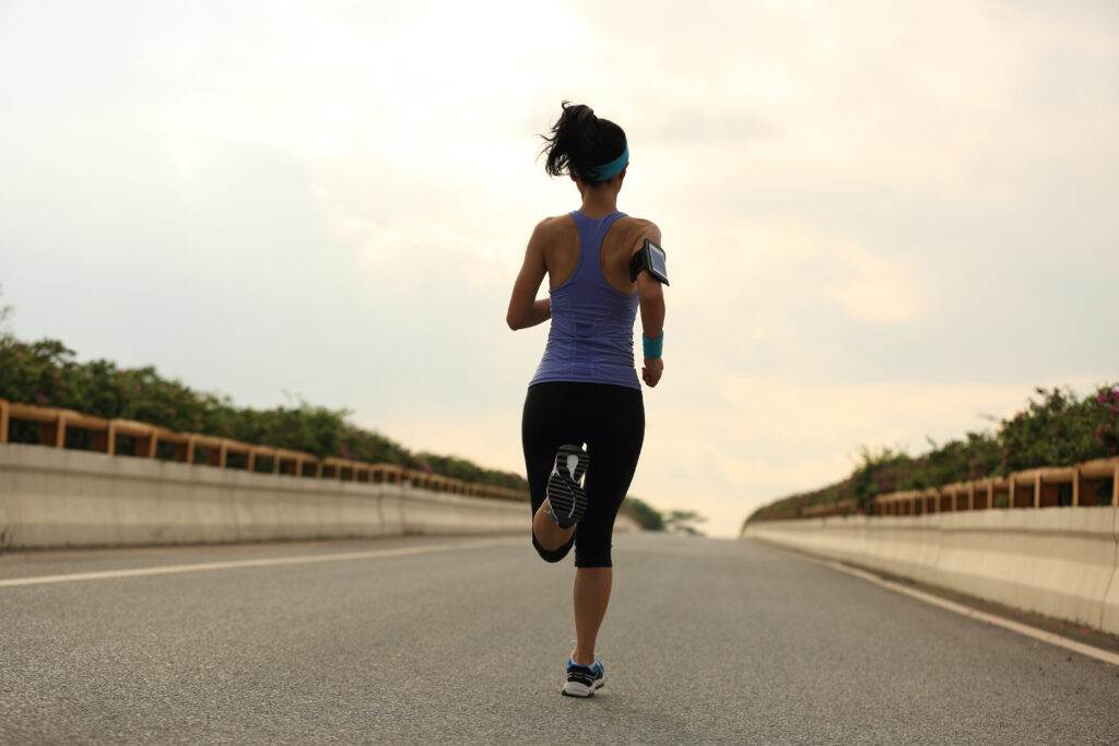 Woman in athletic wear running on an open road, with a fitness armband and headband, surrounded by greenery and a cloudy sky