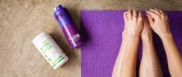 A person sits on a purple yoga mat stretching forward, with a purple water bottle and a container of green superfood powder placed nearby on a brown surface.
