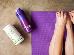 A person sits on a purple yoga mat stretching forward, with a purple water bottle and a container of green superfood powder placed nearby on a brown surface.