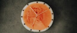 A round slice of watermelon with seeds on the rind arranged like clock numbers and white clock hands, placed on a dark textured surface.