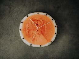 A round slice of watermelon with seeds on the rind arranged like clock numbers and white clock hands, placed on a dark textured surface.