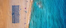 Aerial view of a beach with rows of white umbrellas on golden sand, a few people walking nearby, and turquoise waves gently meeting the shore.