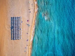 Aerial view of a beach with rows of white umbrellas on golden sand, a few people walking nearby, and turquoise waves gently meeting the shore.