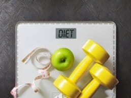 A scale displays the word DIET on its screen, with a green apple, yellow dumbbells, and a measuring tape placed on top, symbolizing healthy living and fitness.